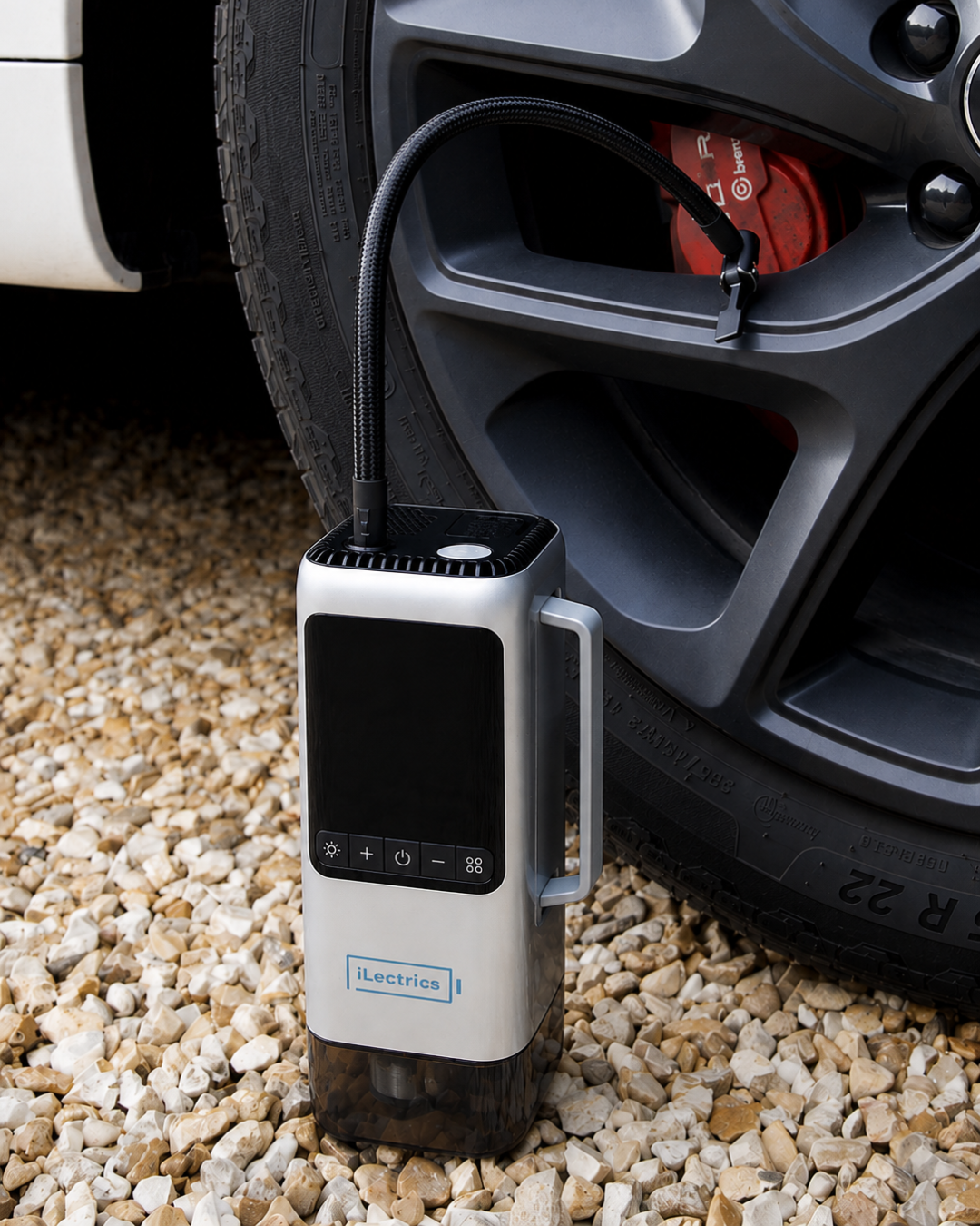 car emergency portable air compressor with a flexible hose, standing on gravel next to a car wheel.